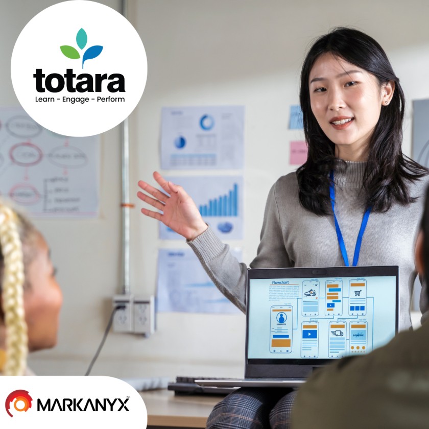 The image features a professional woman leading a presentation in a classroom or office setting. She is gesturing with her hand while holding a laptop that displays a flowchart or infographic, likely related to a training or educational session. The background includes charts and graphs on the wall, suggesting a focus on data and analysis. In the top left corner, the Totara logo is prominently displayed with the text "Totara: Learn - Engage - Perform," highlighting the platform's focus on learning management and performance. In the bottom left corner, the Markanyx Solutions logo is visible, indicating a partnership or collaboration between Markanyx and Totara in delivering educational or training solutions. The overall composition emphasizes themes of digital learning, professional development, and effective collaboration between the two companies.