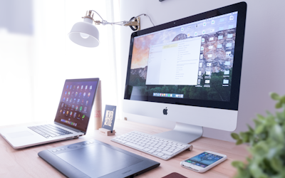 Desktop workspace featuring an iMac, MacBook, drawing tablet, smartphone, and modern lamp, illustrating a creative environment for web design and digital solutions.
